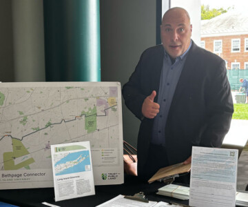 Man in a suit standing behind a table with large map displays at an informational booth about Long Island Greenway and Bethpage Connector, by a window.