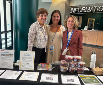 Three women smiling at an information desk in a lobby, with brochures, flyers, and snack cups on the table.