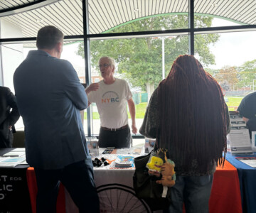 Older man in a white NYBC shirt talks to a man in a suit at a booth with brochures and small giveaways by a windowed wall.